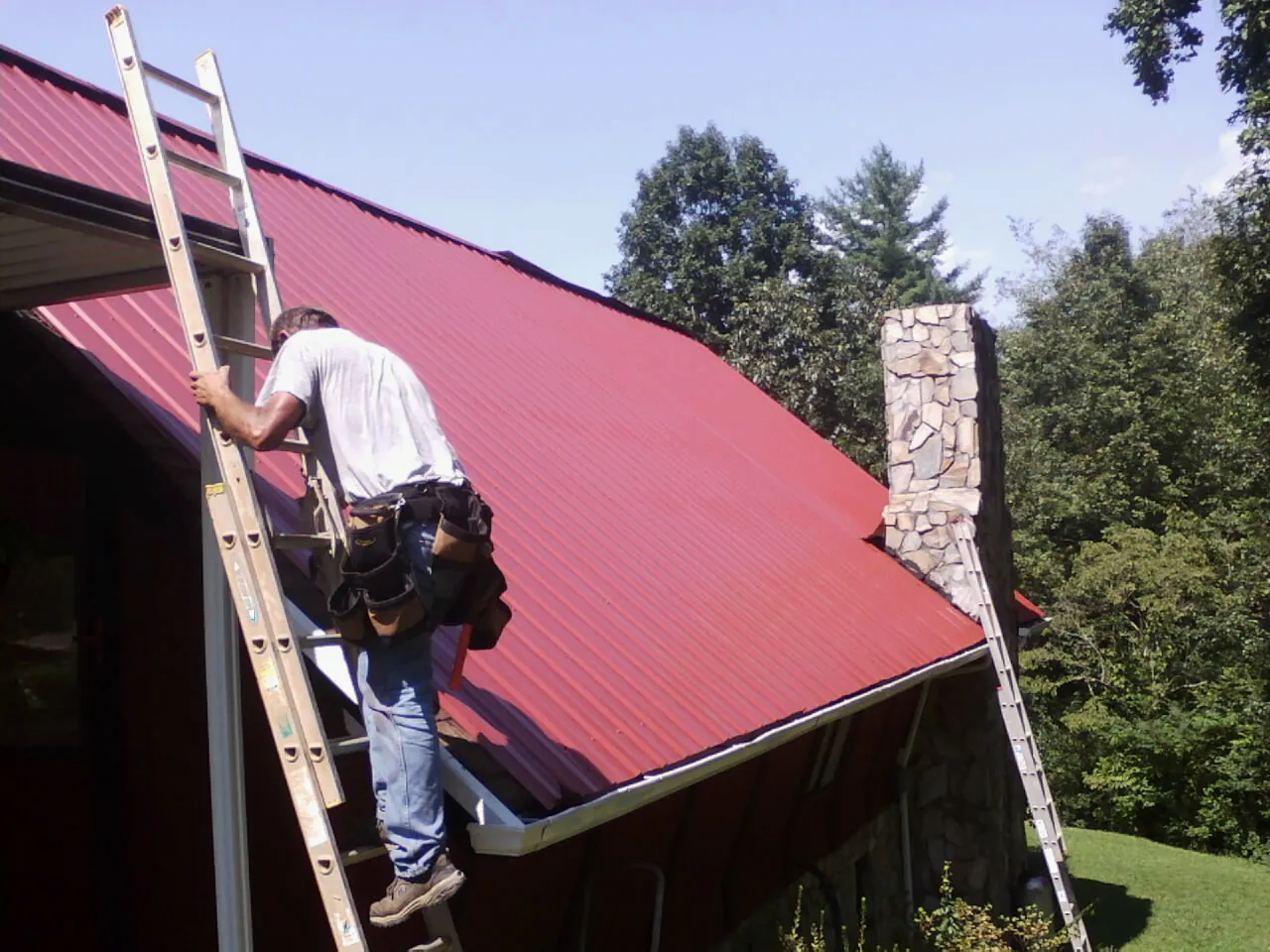 Roofer on a ladder beside a red metal roof and stone chimney — Soffit & Fascia Repair by Roofing Brothers in Weare